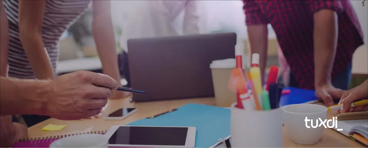 Team collaborating around a table with laptops, a tablet, and creative materials, representing the integration of marketing and web development. Tuxdi logo in the corner.